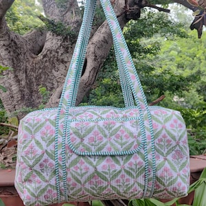 May include: A white and pink floral patterned duffel bag with green trim and straps. The bag is hanging from a tree branch. The floral pattern is repeated across the bag's surface.