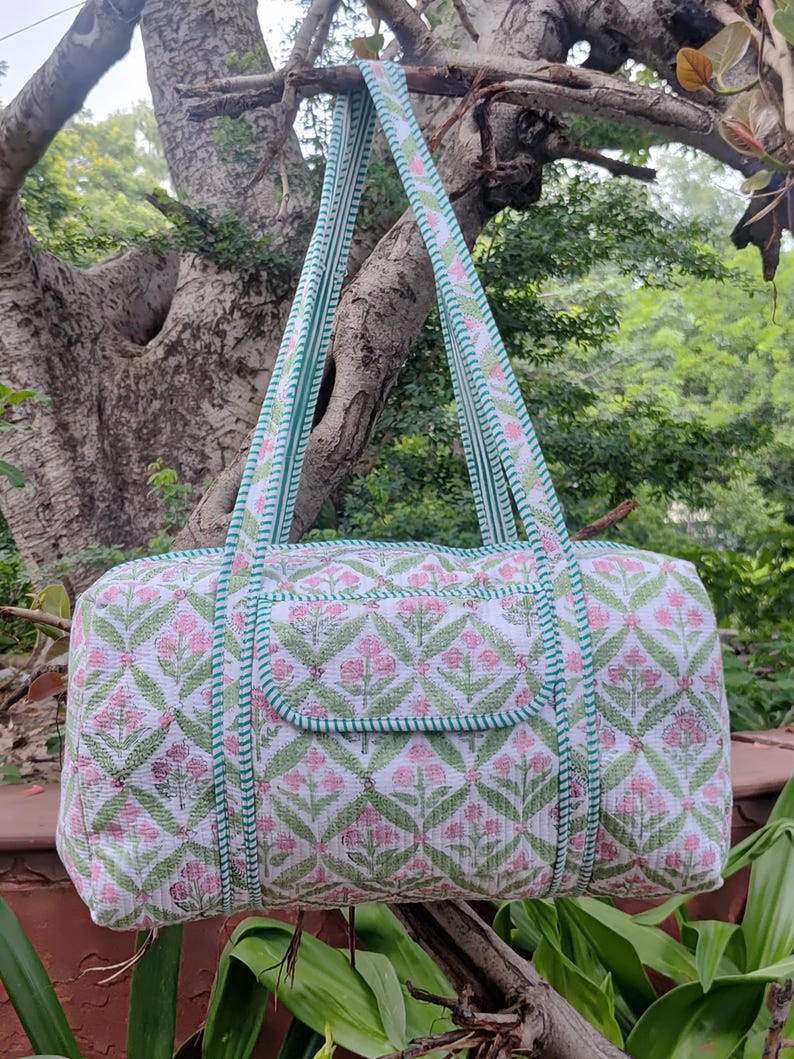 May include: A white and pink floral patterned duffel bag with green trim and straps. The bag is hanging from a tree branch. The floral pattern is repeated across the bag's surface.