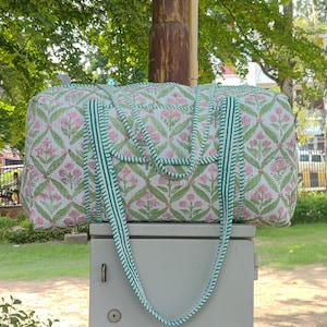 May include: A quilted duffel bag with a floral pattern in pink and green on a white background. The bag has green striped handles and trim. The bag is sitting on a gray box, with a green background of trees and grass.