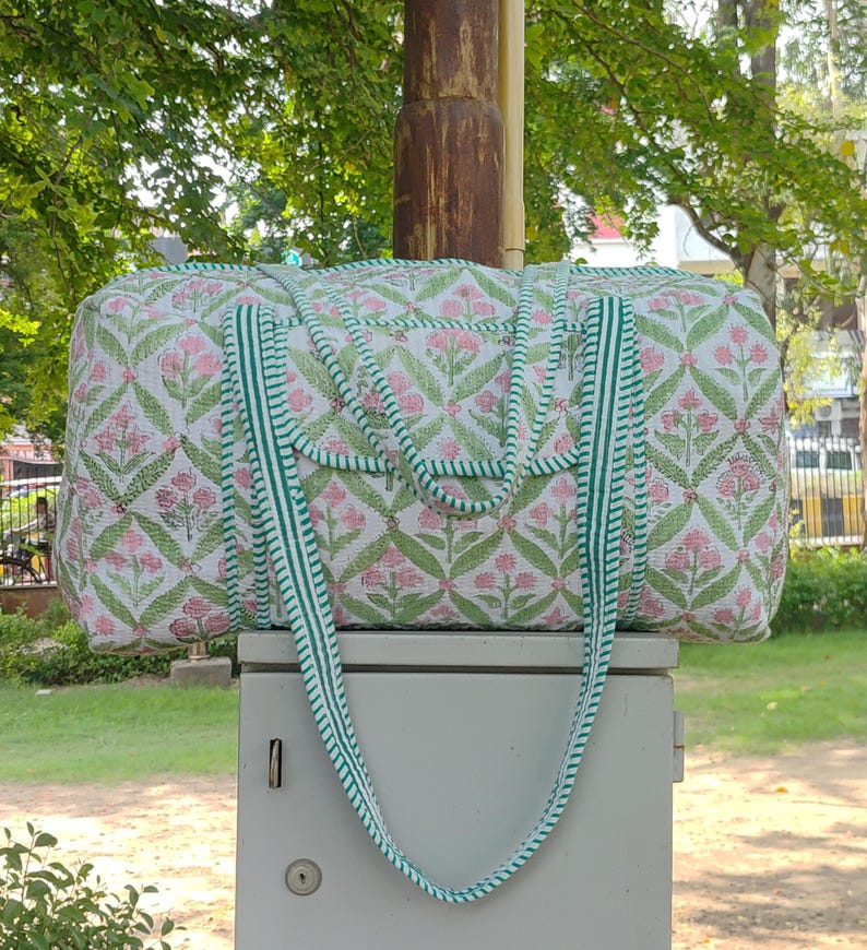 May include: A quilted duffel bag with a floral pattern in pink and green on a white background. The bag has green striped handles and trim. The bag is sitting on a gray box, with a green background of trees and grass.