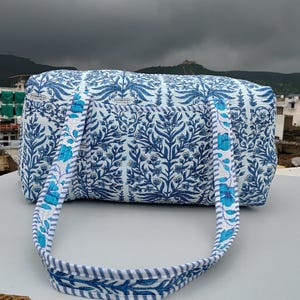 Könnte beinhalten: Eine blaue und weiße Duffle Bag mit floralem Muster und einem gestreiften Riemen. Die Tasche ist aus Baumwolle und hat ein gestepptes Design.