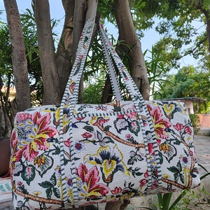 May include: A white quilted duffel bag with floral print featuring pink, yellow, and green flowers. The bag has two long straps and a green and white striped trim.