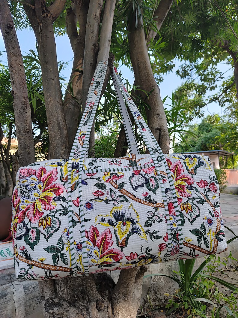 May include: A white quilted duffel bag with floral print featuring pink, yellow, and green flowers. The bag has two long straps and a green and white striped trim.