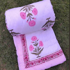 Puede incluir: Edredón floral blanco y rosa con una almohada enrollada a juego. El edredón presenta un diseño floral rosa con tallos y hojas verdes. El borde tiene un patrón de rayas rosas y blancas con un ribete floral. Sobre una superficie de hierba verde.