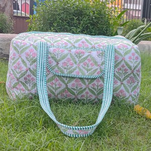 May include: A white and pink floral patterned duffel bag with green accents. The bag has a rectangular shape with a small pocket on the front and green striped handles. The bag is sitting on green grass.