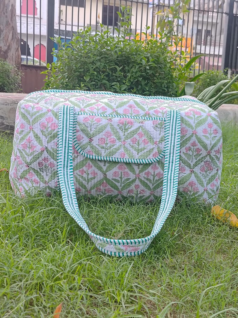 May include: A white and pink floral patterned duffel bag with green accents. The bag has a rectangular shape with a small pocket on the front and green striped handles. The bag is sitting on green grass.
