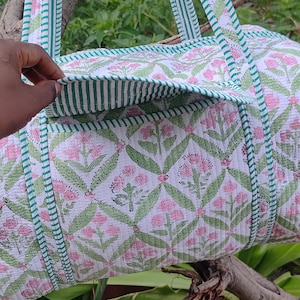 May include: A white quilted duffel bag with a floral pattern in pink and green. The bag has green striped trim and handles. The bag is shown outdoors, resting on a tree branch.