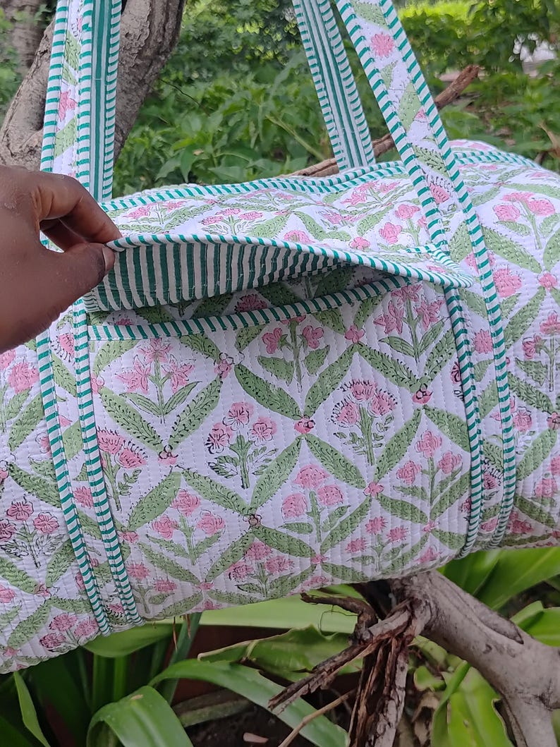 May include: A white quilted duffel bag with a floral pattern in pink and green. The bag has green striped trim and handles. The bag is shown outdoors, resting on a tree branch.