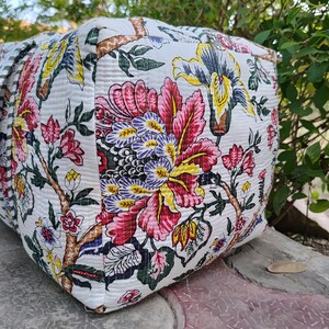 May include: A white fabric tote bag with a floral print in shades of pink, yellow, green, and blue. The bag has a quilted texture and a zipper closure.