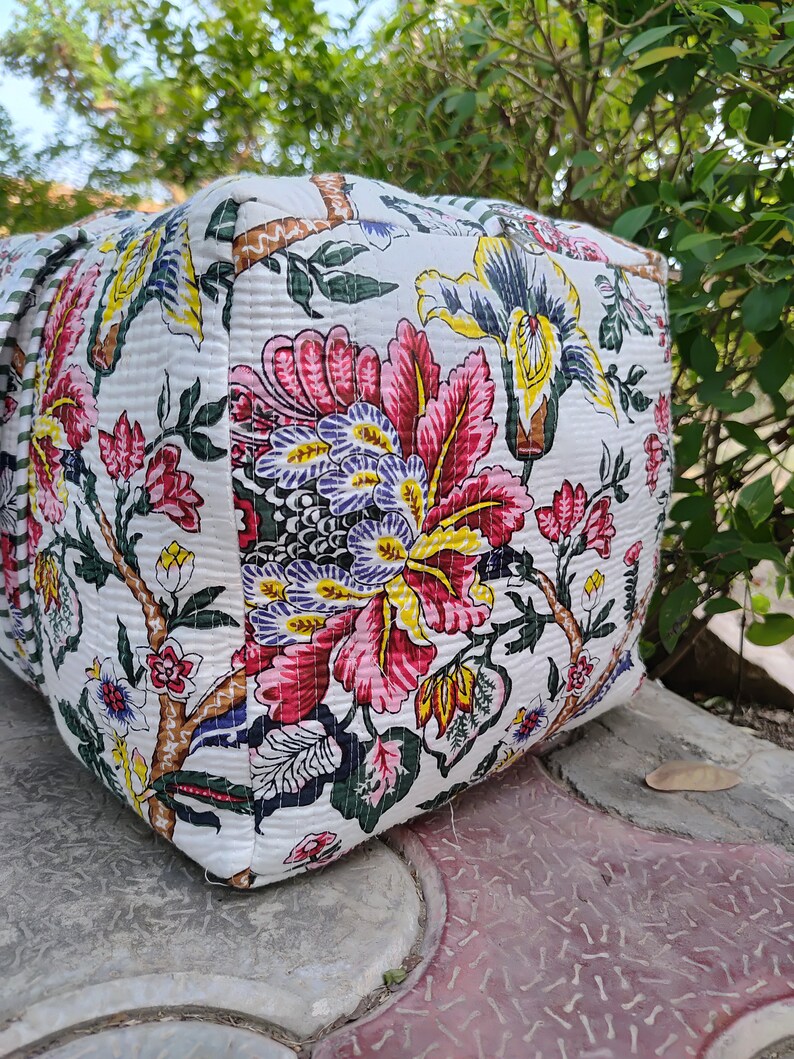 May include: A white fabric tote bag with a floral print in shades of pink, yellow, green, and blue. The bag has a quilted texture and a zipper closure.