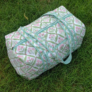 May include: A white and pink floral patterned duffel bag with green accents. The bag has a cylindrical shape with green and white striped handles and trim. The bag is lying on a bed of green grass.