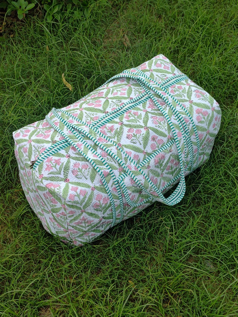 May include: A white and pink floral patterned duffel bag with green accents. The bag has a cylindrical shape with green and white striped handles and trim. The bag is lying on a bed of green grass.