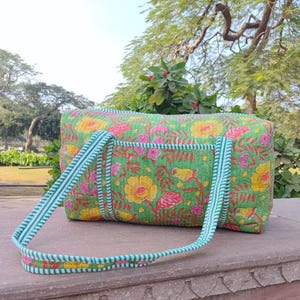 May include: A green quilted duffel bag with a floral print featuring yellow, pink, and red flowers. The bag has white and green striped straps.