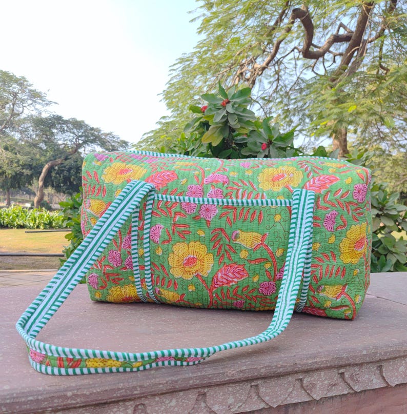 May include: A green quilted duffel bag with a floral print featuring yellow, pink, and red flowers. The bag has white and green striped straps.
