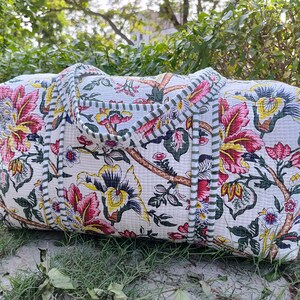 May include: A white quilted duffel bag with a floral print in shades of pink, yellow, and green. The bag has green and white striped trim and two handles.