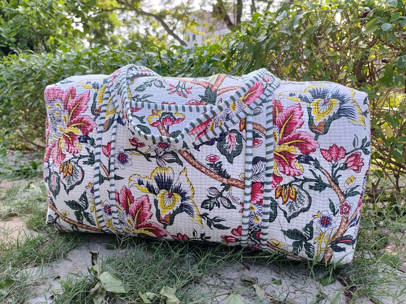May include: A white quilted duffel bag with a floral print in shades of pink, yellow, and green. The bag has green and white striped trim and two handles.