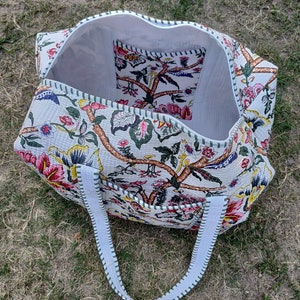 May include: A white quilted tote bag with floral print and green trim. The bag has two white straps and a small interior pocket.