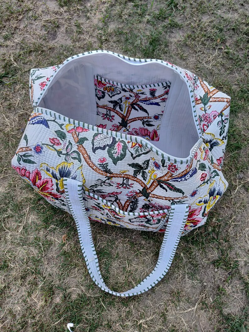 May include: A white quilted tote bag with floral print and green trim. The bag has two white straps and a small interior pocket.