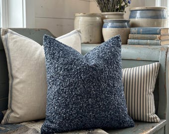 May include: A cozy interior scene featuring three decorative pillows in varying textures and patterns. A large, textured navy blue pillow is centered, flanked by a cream-colored pillow with blue trim and a striped pillow. The pillows rest on a blue bench with a blue and white checkered blanket.