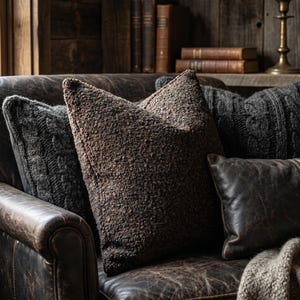 May include: A leather sofa with several cushions in shades of brown and grey. The cushions have different textures, including a bouclé, a cable knit, and a smooth leather. The sofa is in a rustic setting with wooden paneling and books.