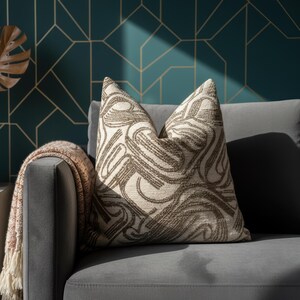 May include: A patterned cushion with a cream base and brown abstract design sits on a grey sofa. A fringed throw and a white vase with gold leaves are also visible. The background features a teal geometric pattern.