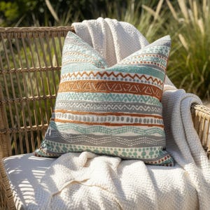 May include: A cushion with a geometric pattern in teal, brown, and grey sits on a white blanket. The cushion is on a wicker chair, with a succulent in a terracotta pot in the background. The cushion's design includes zigzags and dotted lines.