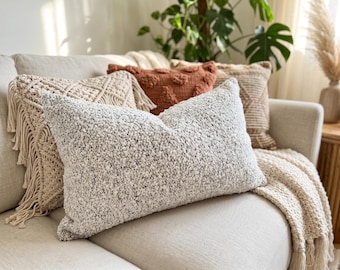 May include: A rectangular throw pillow with a textured, nubby white and black fabric surface. It is positioned in front of other decorative pillows, including a cream macrame pillow with fringe and a solid rust-colored pillow.
