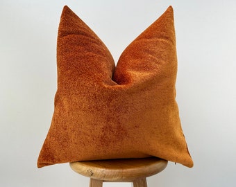 May include: A burnt orange velvet pillow with a soft, textured surface. The pillow is sitting on a wooden stool.