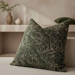 May include: A square decorative pillow with a dark green and beige palm leaf pattern. The pillow is angled, showcasing the detailed print and texture. The pillow is on a neutral-toned surface, suggesting a home decor setting.
