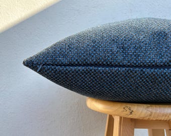 May include: A blue woven textured pillow with a zipper closure. The pillow is resting on a small wooden stool. The pillow's fabric has a mix of blue and black threads.