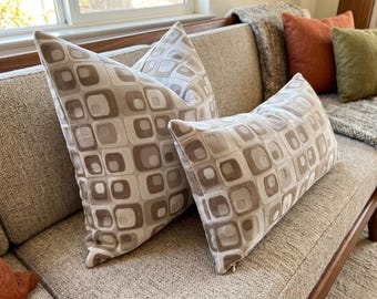 May include: Two throw pillows featuring a repeating pattern of rounded squares in shades of beige, tan, and cream. The square pillow is positioned behind a rectangular lumbar pillow, both displaying the same geometric design on a soft fabric.