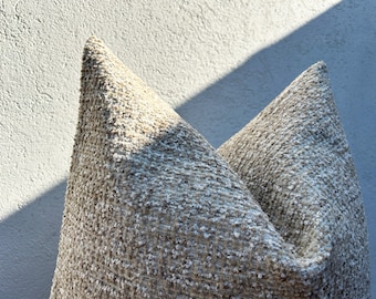 May include: Close-up of two textured throw pillows. The pillows are a neutral color with a speckled pattern. The fabric appears to be a woven material. The pillows are in a V-shape, with the tips pointing upwards.