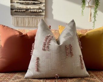 May include: Three decorative throw pillows in shades of orange, gold, and beige. The center pillow has a textured pattern with pink fringe accents. The pillows are arranged on a patterned sofa, creating a cozy and stylish home decor scene.