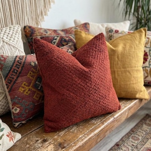 May include: A selection of decorative cushions in various colours and patterns. The cushions include a textured rust-coloured cushion, a mustard yellow cushion, and cushions with patterned designs. The cushions are arranged on a wooden bench.