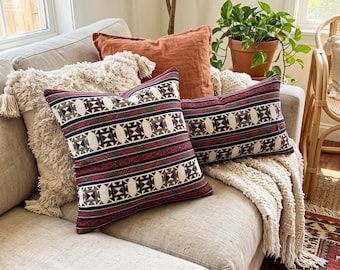 May include: Two square and rectangular throw pillows featuring a geometric pattern in black, white, and deep red. The pillows are displayed alongside a textured cream-colored pillow with tassels and a solid burnt orange pillow.