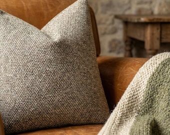 May include: A textured, neutral-toned throw pillow with a brown leather armchair. The pillow has a woven appearance, with a mix of light and dark threads. A folded green and white blanket is draped over the arm of the chair. The background includes a stone wall and wooden table.