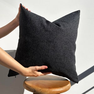 May include: A dark gray throw pillow with a textured fabric. The pillow is being held by a hand and is sitting on a wooden stool.