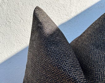 May include: Close-up of two decorative pillows with a dark brown and black herringbone pattern. The pillows are angled towards the viewer, showcasing the texture and design. The pillows are against a white textured wall.