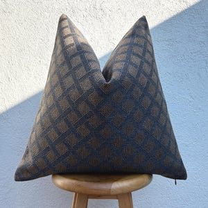 May include: A decorative throw pillow with a geometric pattern. The pillow features a repeating diamond design in shades of brown and navy blue. The pillow is placed on a small wooden stool against a textured white and blue wall.