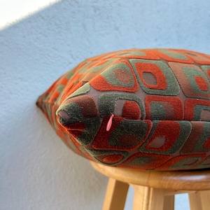 May include: A decorative pillow with a geometric pattern in shades of orange, green, and brown. The pillow is placed on a small wooden stool. The pillow's design features squares and rectangles, creating a retro aesthetic.