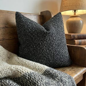 May include: A dark gray textured pillow with a pointed top rests on a wooden bench. A knitted blanket with gray and cream stripes is draped below the pillow. A lamp and books are in the background, creating a cozy, homey scene.