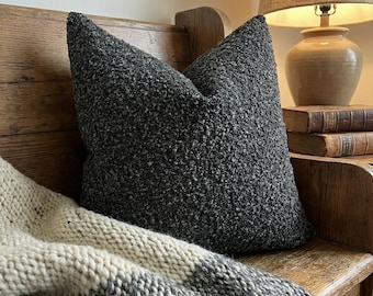 May include: A dark gray textured pillow with a pointed top rests on a wooden bench. A knitted blanket with gray and cream stripes is draped below the pillow. A lamp and books are in the background, creating a cozy, homey scene.