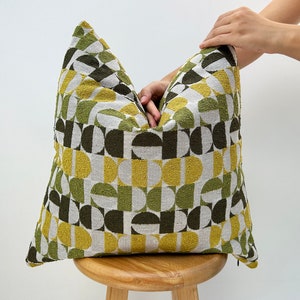 May include: A green, yellow and white geometric patterned cushion with a zip closure. The cushion is sitting on a wooden stool.