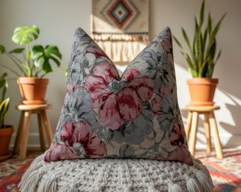 May include: A decorative throw pillow with a floral pattern in shades of gray and pink. The pillow is placed on a white, textured pouf. Potted plants and a small wooden stool are in the background, creating a cozy, homey atmosphere.