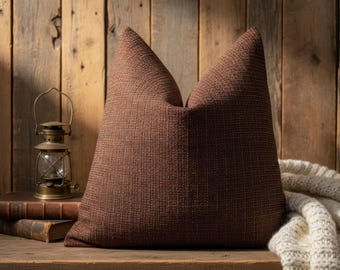 May include: A large, brown textured pillow with a woven appearance. The pillow is placed on a wooden surface, with a vintage lantern and old books in the background. A cream-colored knitted blanket is partially visible.