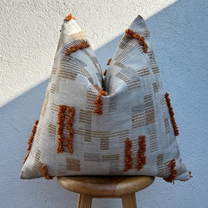 May include: A decorative cushion with a neutral colour scheme and a geometric pattern. The cushion features a light beige background with brown and orange accents. The design includes rectangular shapes and textured, fringed details. The cushion is placed on a wooden stool.