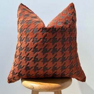 May include: A brown and black patterned throw pillow with a houndstooth design. The pillow is sitting on a wooden stool.