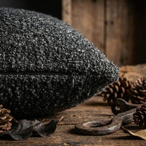 May include: Close-up of a textured, dark gray decorative pillow with a zipper closure. The pillow has a nubby, bouclé-like fabric. The background includes pine cones and a rustic wooden surface, suggesting a cozy, natural aesthetic.