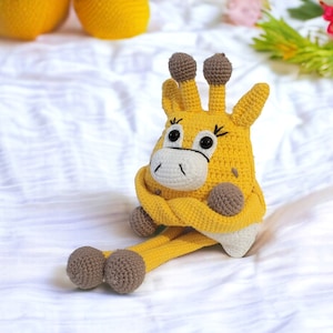 Puede incluir: Un juguete de jirafa de crochet amarillo con patas y orejas marrones. La jirafa está sentada sobre una superficie blanca.