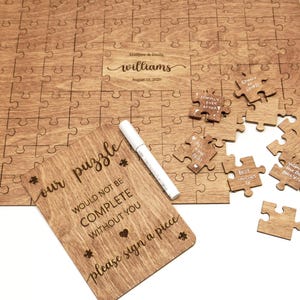 May include: A wooden jigsaw puzzle guest book with engraved text reading "Matthew & Emily Williams August 17, 2025" and a separate wooden sign reading "our puzzle WOULD NOT BE COMPLETE WITHOUT YOU please sign a piece" with a white marker.